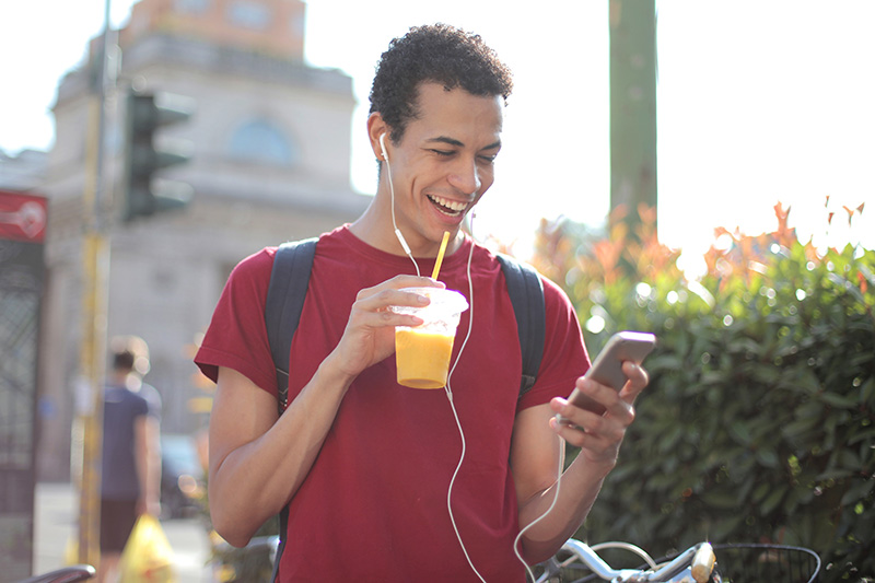a man standing on the sidewalk looking at his smartphone while laughing and drinking juice